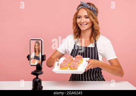 Jeune femme adulte blogueuse montrant des gâteaux cuits au four et le tournage sur vidéo, enregistrement vidéo pour la chaîne alimentaire, femme pâtissière vlogging avec mobile phon Banque D'Images