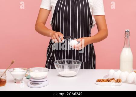 Image rognée d'une femme boulangère sans visage faisant de la pâte, conduisant l'œuf à la farine pour faire de la pâte, portant un tablier rayé noir, ingrédients pour la boulangerie. Intérieur Banque D'Images