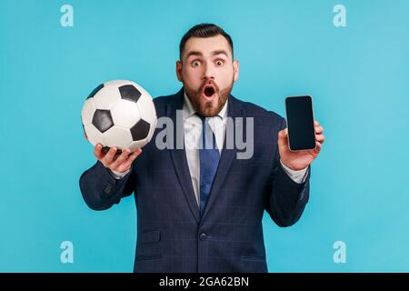 Stupéfait homme choqué avec la barbe dans le style officiel costume tenant et montrant le smartphone avec écran noir et ballon de football, pariant sur le football. Studio intérieur Banque D'Images