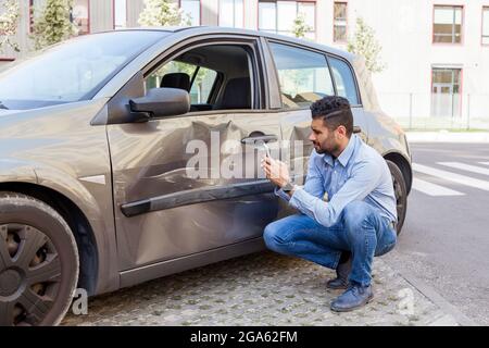 Portrait de brunette homme portant un Jean et une chemise bleue ne pas faire face à la conduite, a endommagé sa porte de voiture, des bosses et des rayures sur le corps de voiture, faisant des photos de Banque D'Images