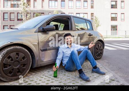 Jeune adulte contrarié conducteur homme portant un Jean et chemise bleue assis près d'une voiture de collision, accident de collision dans la route de la ville, ne conduisez pas ivre, porte de voiture endommagée Banque D'Images