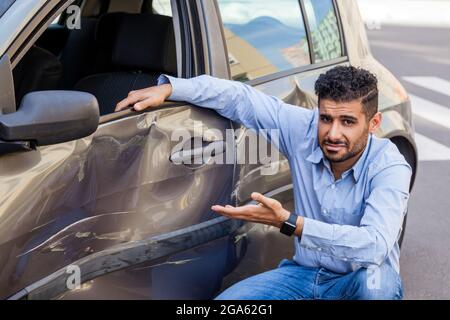 Portrait de beau barbu homme portant un Jean et une chemise assis près de la voiture et montrant avec les mains des bosses et des rayures sur la porte de son automobile, d Banque D'Images