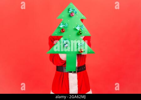 Inconnu homme portant le costume du père noël cachant son visage derrière un beau papier vert décoré arbre de Noël, surprise de la Saint-Sylvestre. Intérieur s Banque D'Images