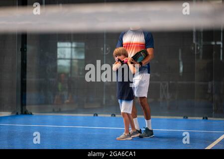 Surveiller l'enseignement de la classe de padel à l'enfant, son étudiant - formateur enseigne à petit garçon comment jouer de padel sur un court de tennis intérieur Banque D'Images