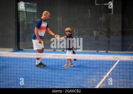 Surveiller l'enseignement de la classe de padel à l'enfant, son étudiant - formateur enseigne à petit garçon comment jouer de padel sur un court de tennis intérieur Banque D'Images