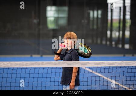 Surveiller l'enseignement de la classe de padel à l'enfant, son étudiant - formateur enseigne à petit garçon comment jouer de padel sur un court de tennis intérieur Banque D'Images