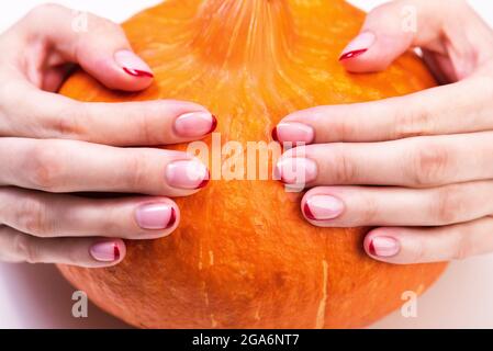 Mains de femmes avec une manucure française d'automne tendance avec des bouts rouges au bord libre de l'ongle. Citrouille orange sur fond blanc. Courge orange pour TH Banque D'Images