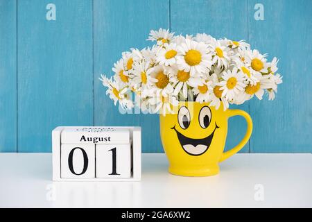 1er août date sur le calendrier et bouquet de camomille en tasse jaune avec visage drôle contre mur bleu. Concept du mois d'été Hello August. Banque D'Images