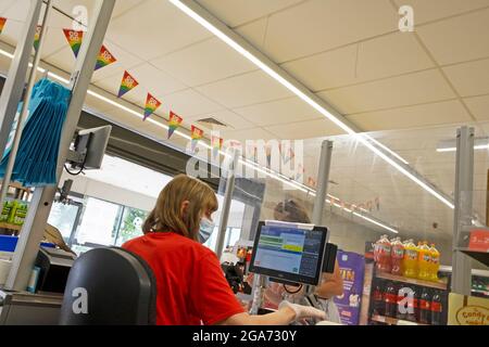 Vue arrière d'une femme plus âgée, employée assise au comptoir de caisse iat, écran portant un masque facial à la caisse, dans le supermarché Co Op UK KATHY DEWITT Banque D'Images