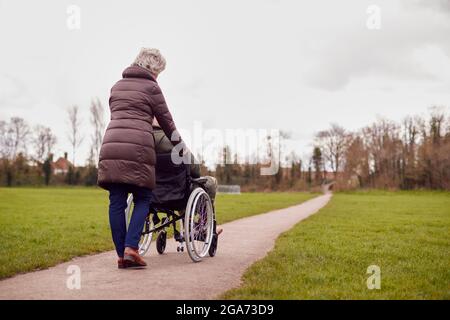 Vue arrière d'une femme âgée poussant un homme en fauteuil roulant à l'extérieur dans le parc d'automne ou d'hiver Banque D'Images