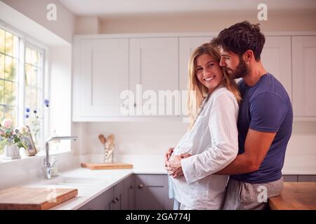 Couple avec homme embrassant femme enceinte portant un pyjama debout dans la cuisine tenant Bump Banque D'Images