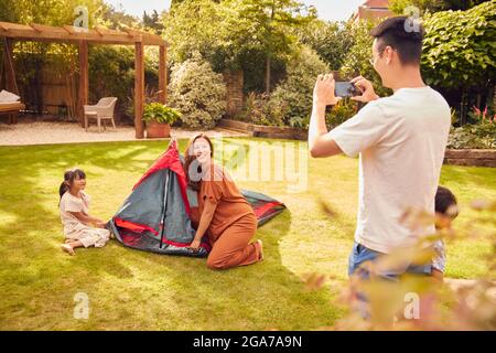 Père asiatique prenant des photos sur téléphone portable comme famille de tente d'installation dans le jardin à la maison Banque D'Images