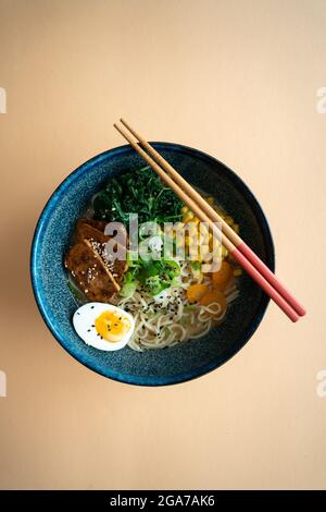 Vue de dessus d'un bol de soupe végétarienne de nouilles ramen avec tofu, œuf dur, oignon de printemps, maïs, bouillon sur fond orange Banque D'Images