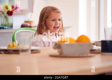 Une jeune fille souriante portant un pyjama assis à la table dans la cuisine à la maison en attendant le petit-déjeuner Banque D'Images