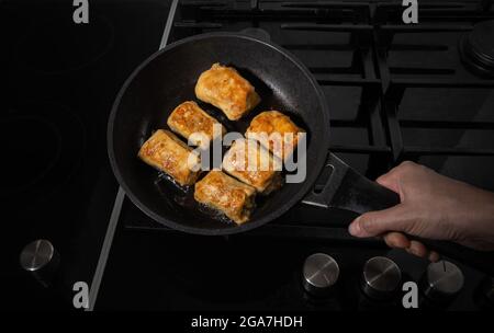 Cuisson de petits pains de chou farcis faits maison. Peu de petits pains sont frits sur une poêle et sur une cuisinière noire. Poêle à frire est à la main de la femme. Huile frite Banque D'Images