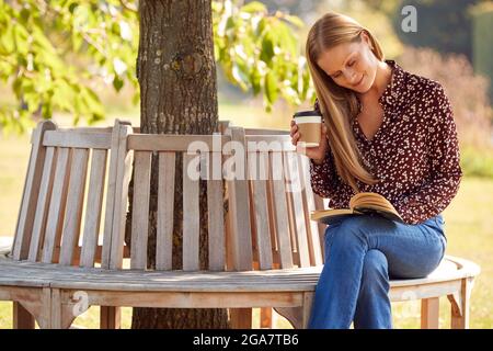 Femme d'âge mûr se détendant assis sur un banc de parc sous un livre de lecture d'arbre avec un café Takeaway Banque D'Images