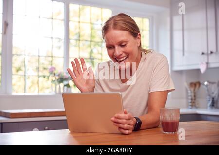 Femme mature à la maison dans la cuisine agitant comme elle fait appel vidéo sur la tablette numérique Banque D'Images