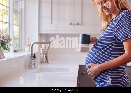 Gros plan de la femme enceinte debout près de la fenêtre de la cuisine touchant l'estomac se relaxant avec une boisson chaude Banque D'Images