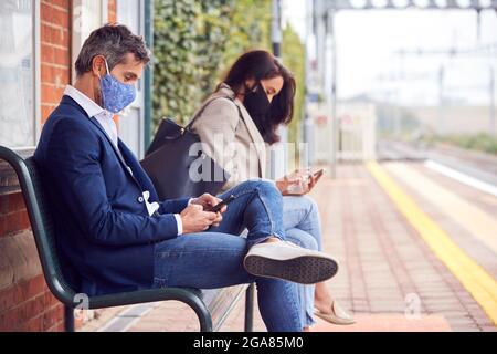Les voyageurs d'affaires se trouvant sur la plate-forme ferroviaire avec des téléphones mobiles portant des masques faciaux pendant une pandémie Banque D'Images