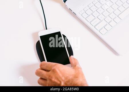 Main avec un smartphone mobile blanc, charge sur une base de chargeur sans fil à côté d'un ordinateur portable sur une table de travail blanche Banque D'Images