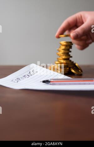 Main mettant la pièce de monnaie dorée sur une colonne de pièce de monnaie dorée avec une liste de gains sur une feuille de papier et un stylo noir sur une table en bois Banque D'Images