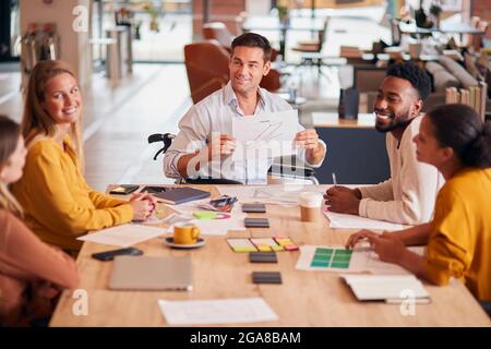 Un homme d'affaires en fauteuil roulant donne une présentation aux collègues autour de la table dans le bureau de plan ouvert moderne Banque D'Images