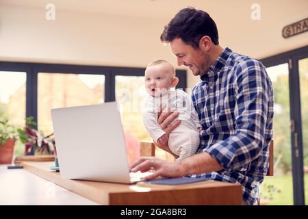 Père travaillant utilisant un ordinateur portable à la maison tout en tenant bébé souriant sur le genou Banque D'Images