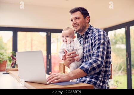 Père travaillant utilisant un ordinateur portable à la maison tout en tenant bébé souriant sur le genou Banque D'Images
