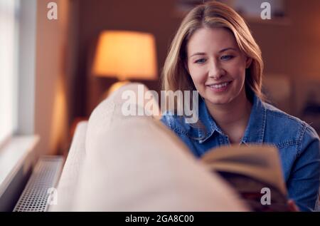 Femme à la maison détente et lecture livre assis sur un canapé le soir Banque D'Images