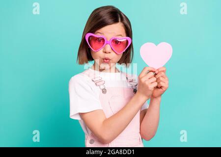 Photo d'une écolière très impressionnée habillé rose général lunettes tenant le coeur de papier isolé couleur turquoise fond Banque D'Images