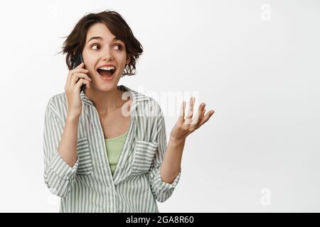Bonne fille moderne parlant sur téléphone mobile, souriant et regardant surpris, debout sur fond blanc Banque D'Images