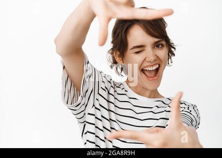 Portrait en gros plan d'une jeune femme heureuse regardant à travers les cadres de la main, recherchant la perspective de prise de vue, s'inspirant, se tenant sur du blanc Banque D'Images