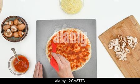 Garniture de pâte à pizza avec de la sauce tomate, préparation en gros plan, plat. Recette de pizza maison à la mozzarella et aux champignons étape par étape Banque D'Images