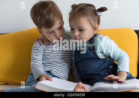 Deux enfants avant-garçonnets lisent un grand livre intéressant de contes de fées sur un lit jaune. Frères et sœurs petits jumeaux lecteur ont plaisir, heureux enfant sur Banque D'Images