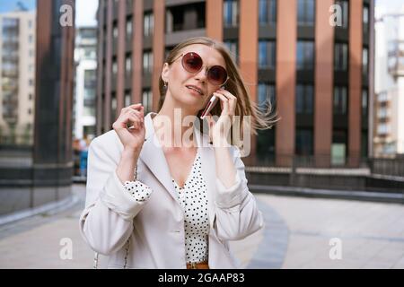Femme d'affaires avec téléphone près du bureau. Portrait de belle fille souriante dans des vêtements de bureau légers à la mode, parlant au téléphone, debout à l'extérieur dans des lunettes de soleil. Communications téléphoniques. Banque D'Images