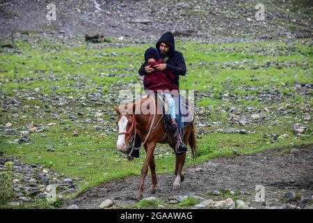 Un homme couvre son enfant avec un tissu tandis qu'ils aiment une promenade à cheval pendant une pluie à Sonamarg, à environ 100 km de Srinagar. Le Département météorologique a prédit jeudi une augmentation des précipitations au Jammu-et-Cachemire, et a également émis une alerte météorologique demandant aux personnes vivant dans les zones vulnérables d'être prudentes. Sept personnes ont été trouvées mortes et 17 autres ont été sauvées dans un état blessé après que le village éloigné a été frappé par l'explosion de nuages dans les premières heures de mercredi, endommageant 21 maisons, des hangars à vache, un dépôt de ration, un pont et une mosquée. Banque D'Images