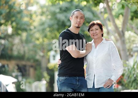 Portrait d'une femme asiatique mûre souriant à l'appareil photo tout en se tenant debout à l'extérieur avec son fils aîné Banque D'Images