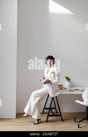 Femme d'affaires en costume blanc assise sur un bureau dans un bureau moderne. Banque D'Images