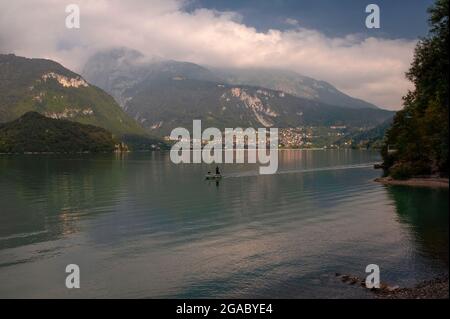 Un homme se dresse dans son petit bateau à moteur, regardant de l'autre côté de la rive, alors qu'il traverse la surface placide du «plus beau lac d'Italie», le réservoir alpin Lago di Molveno dans le Trentin-Haut-Adige, Sur fond de la ville de Molveno, située au-dessous des sommets des Dolomites de Brenta, enveloppés de nuages. La population de Molveno, qui compte environ 1,100 000 habitants, est gonflée en été par de nombreux visiteurs attirés par la beauté naturelle du lac, ses plages, son eau propre et ses activités telles que la voile, la planche à voile, la pêche, le canoë-kayak et le VTT. Banque D'Images