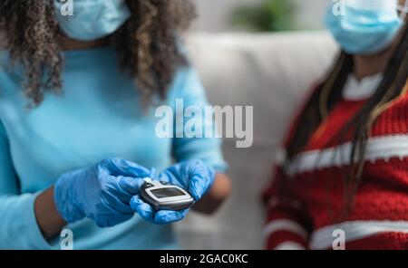 Jeune infirmière vérifiant le test de glycémie pour le diabète à un patient âgé tout en portant des gants chirurgicaux et un masque facial - concept de soins de santé Banque D'Images