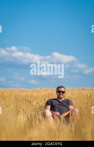 Un jeune homme portant des lunettes de soleil est assis au milieu d'un champ triticale, un jour d'été ensoleillé. Concept de l'agriculture et des affaires. Photo verticale Banque D'Images