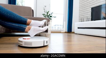 Aspirateur robot qui nettoie la pièce, jambes relevées. Concept de maison intelligente Banque D'Images