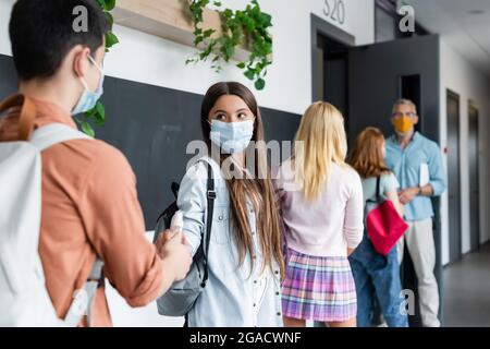 adolescent donnant la main antiseptique à la fille dans le masque médical près de camarades de classe flous et professeur Banque D'Images
