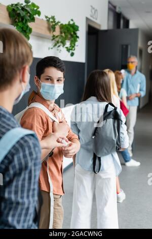 garçon dans un masque médical prenant l'assainisseur pour les mains d'un ami tout en se tenant dans la ligne dans le couloir de l'école Banque D'Images