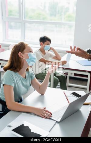 Écolière dans un masque médical parlant à un professeur avec une tablette numérique pendant la leçon Banque D'Images