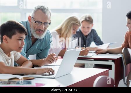 Un professeur mature se dirige vers un ordinateur portable près d'un élève asiatique en classe Banque D'Images