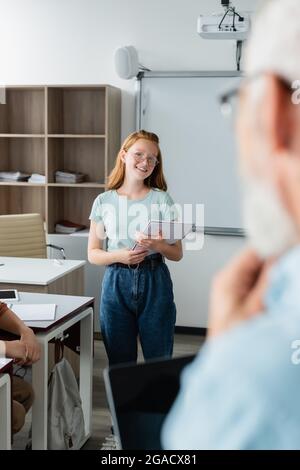 Une écolière souriante tenant un carnet près d'un professeur flou dans la salle de classe Banque D'Images