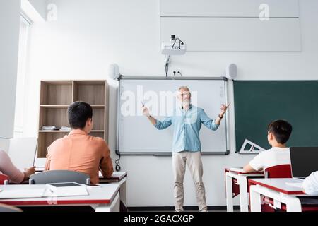 vue arrière des camarades de classe près de l'enseignant parlant et pointant avec les doigts pendant la leçon Banque D'Images