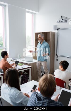 professeur souriant parlant et faisant des gestes près des élèves avec des gadgets dans la salle de classe Banque D'Images