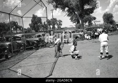 Dans les années 1950, les automobiles historiques de l'époque garées par un terrain de balle ou sandlot, alors que les hommes locaux ont un match de base-ball dans le centre-ouest américain, avec la batte, le attrape et l'arbitre tous prêts pour que le ballon soit incliné. Banque D'Images